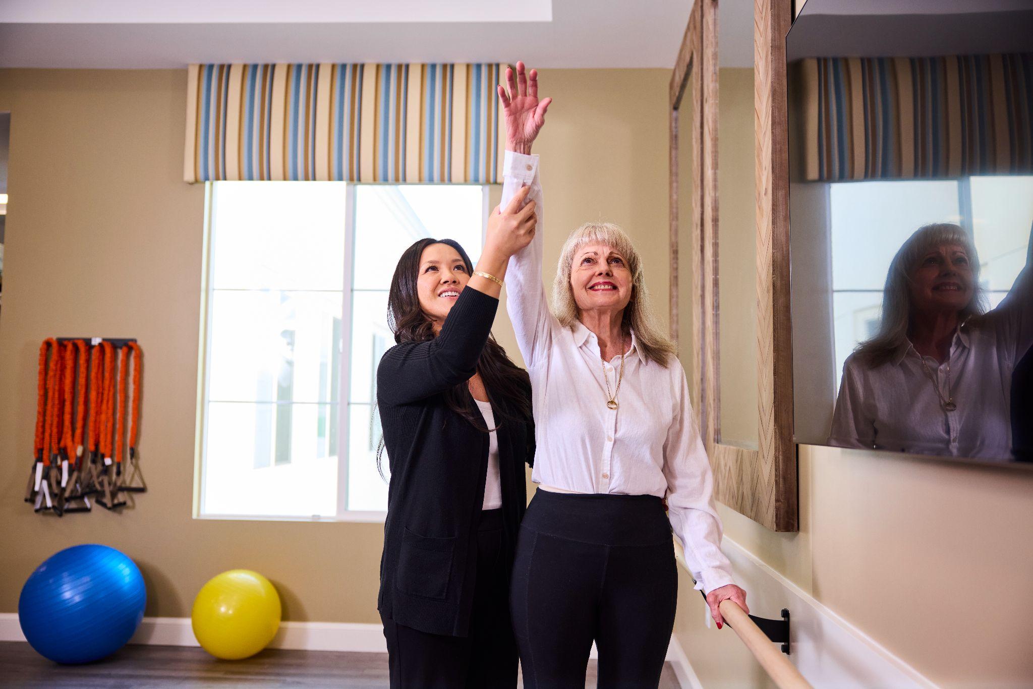 A caregiver assists a senior woman as she reaches her arm overhead during a physical therapy session focused on functional fitness.